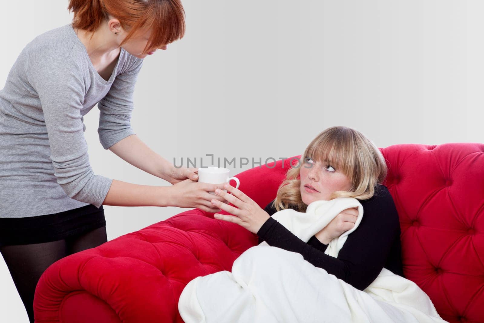 young beautiful blond and red haired girls has fever and get a tea on red sofa in front of grey background