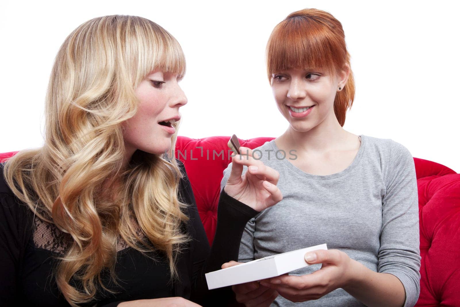young beautiful blond and red haired girls eating chocolate on red sofa in front of white background