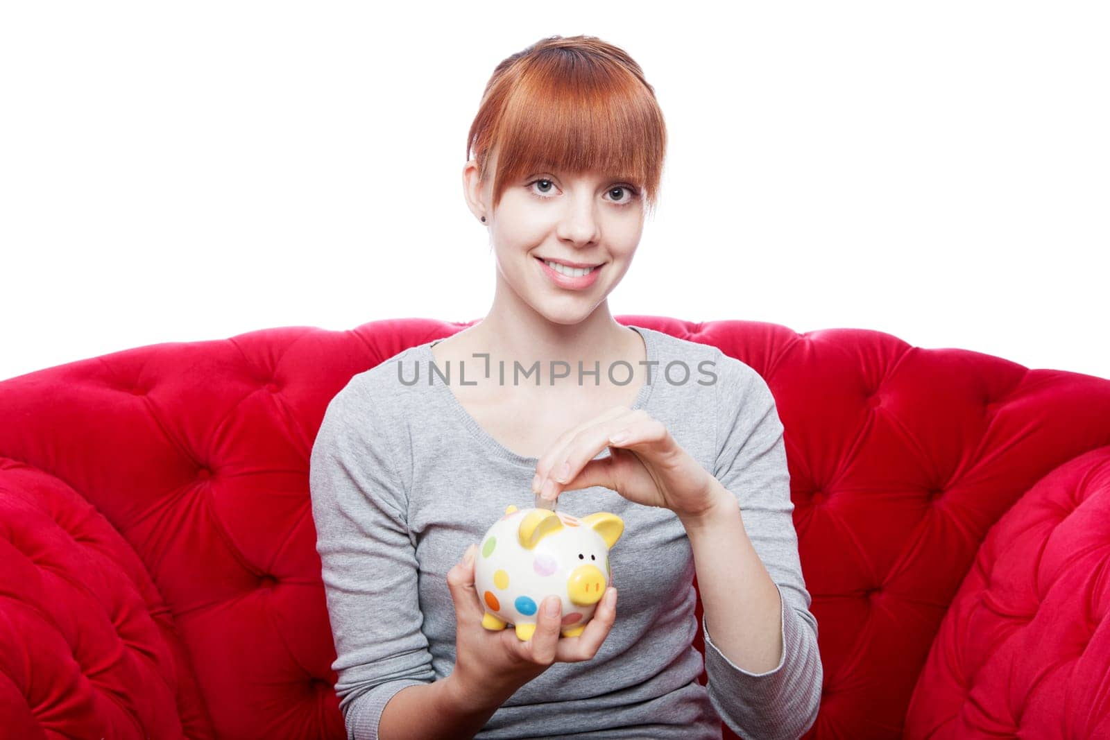 young beautiful red haired girl put coin in piggybank on red sofa in front of white background