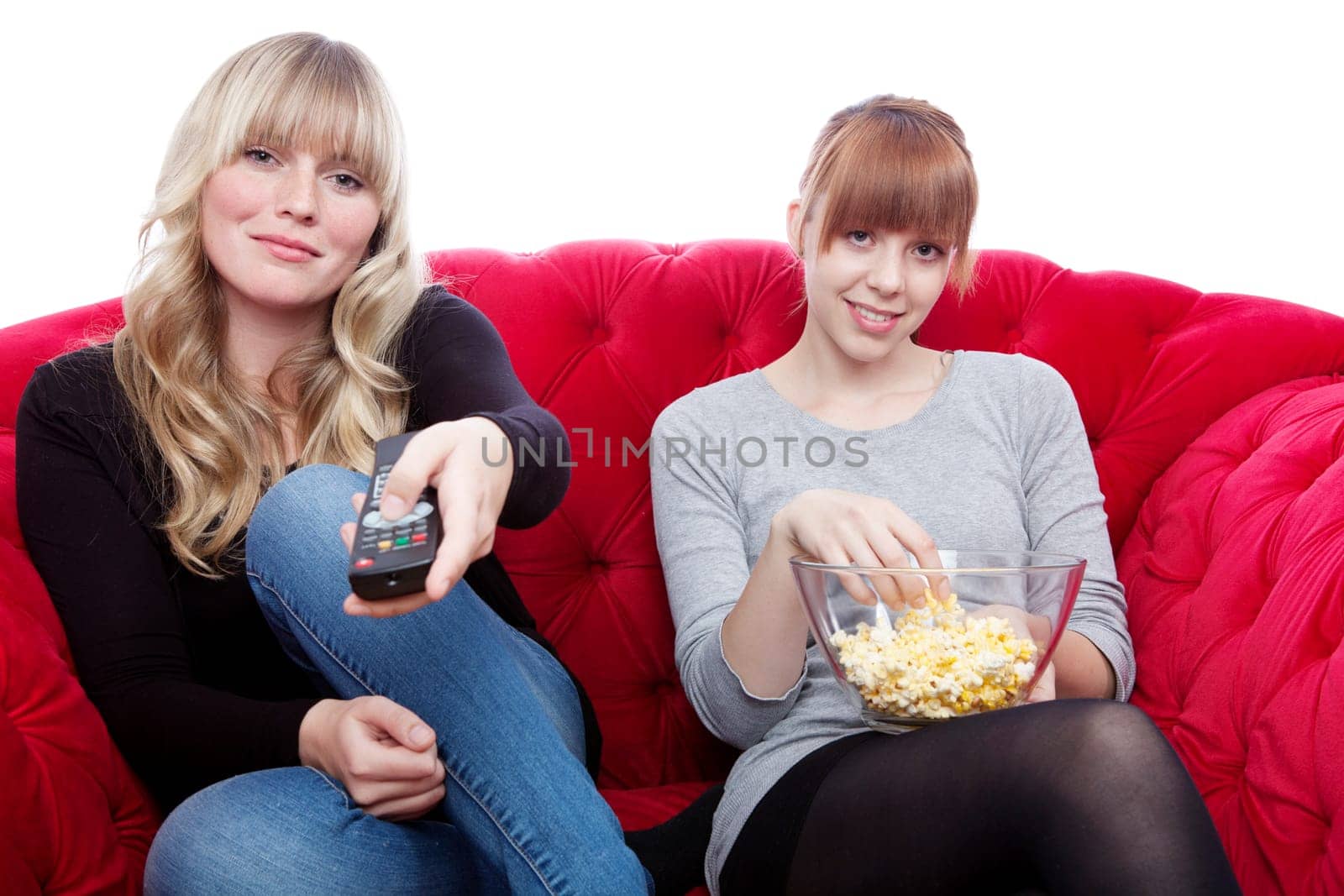 young beautiful blond and red haired girls on red sofa with remote control and popcorn in front of white background