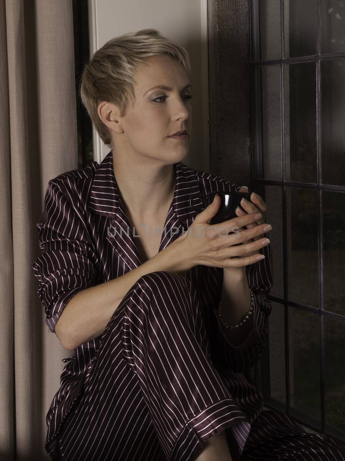Beautiful blond girl by window drinking a mug of tea or coffee.