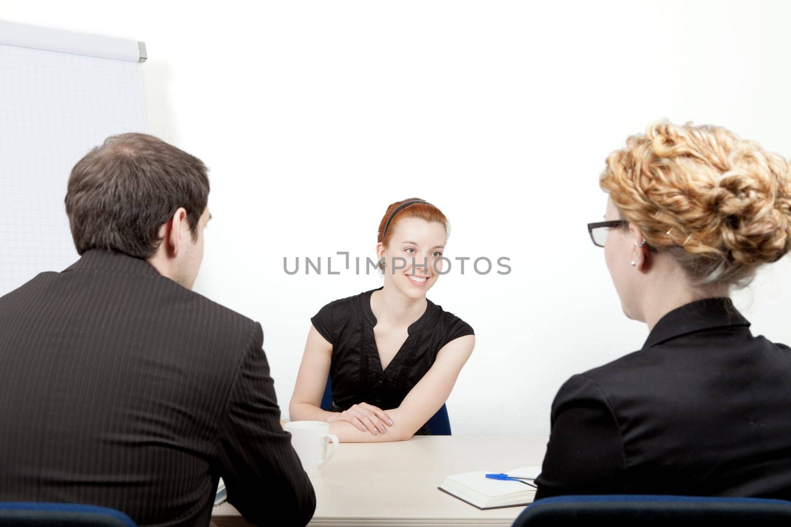 Young woman being interviewed for a job smiling as she answers questions from the two corporate interviewers, a man and woman, who are sitting with their backs to the camera