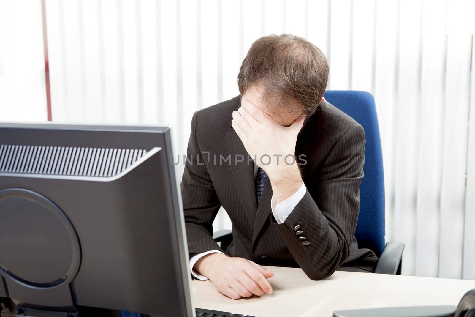 Despairing businessman sitting at his desk in his office resting his forehead in his hands as he considers the hopelessness of the situation