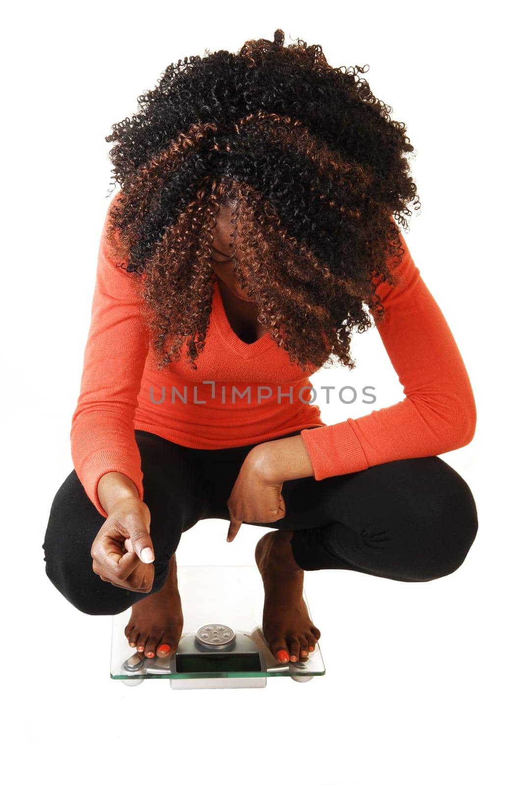A lovely black teen girl crouching on a scale, looking on her little weight, for white background in an orange sweater and black tights.