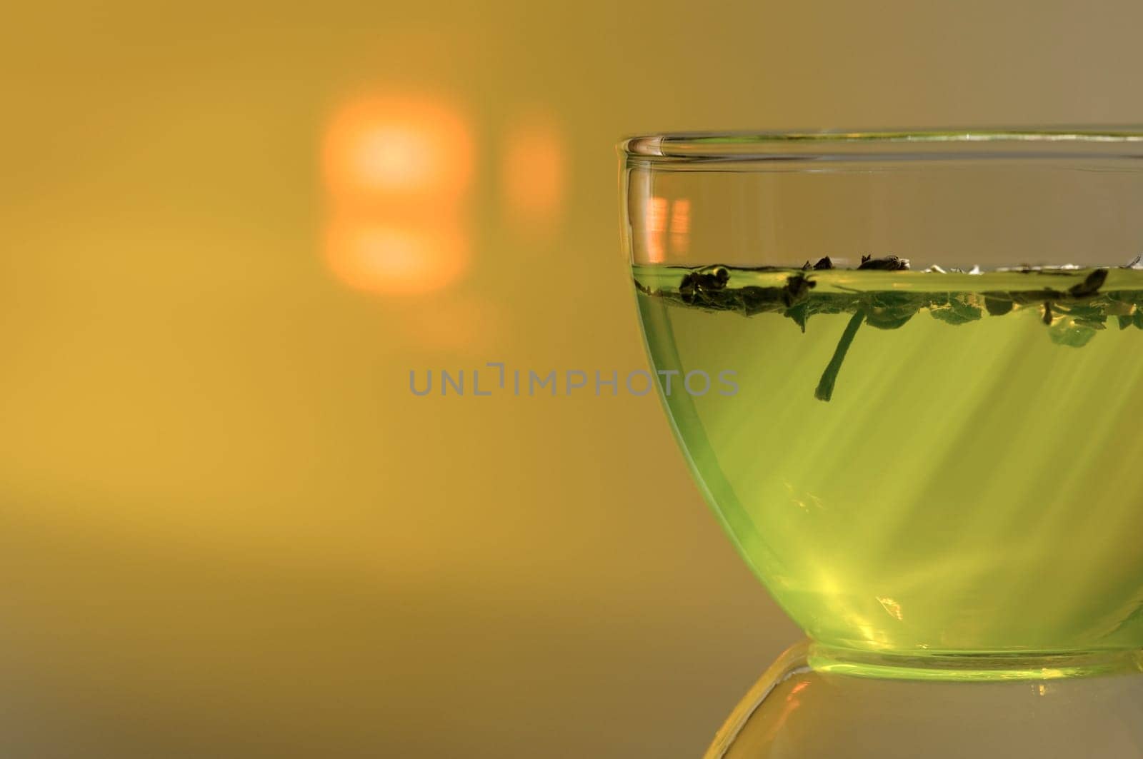 Close-up studio shot of a filled tea glass with tea leaves floating above and oblique light rays in the foreground with background projection of a sun-like backlight