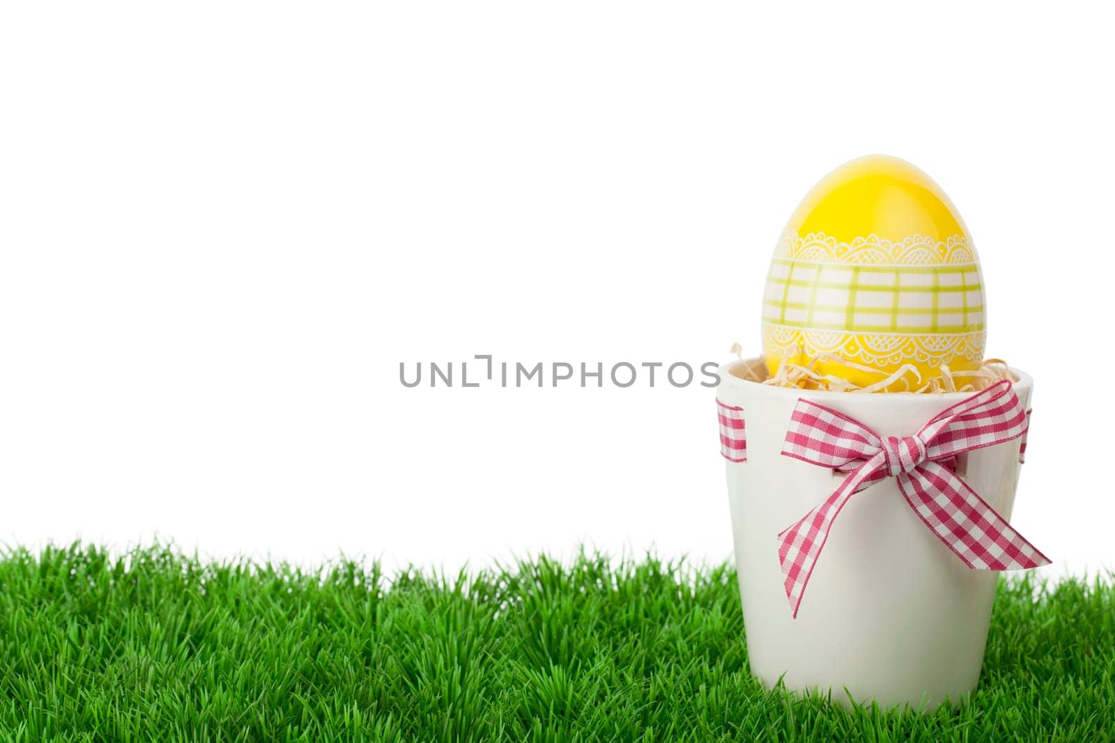 Pretty Easter gift with a decorative yellow Easter Egg inside a flower pot tied with a rustic red and white checked ribbon and bow standing on green grass against a white background with copyspace