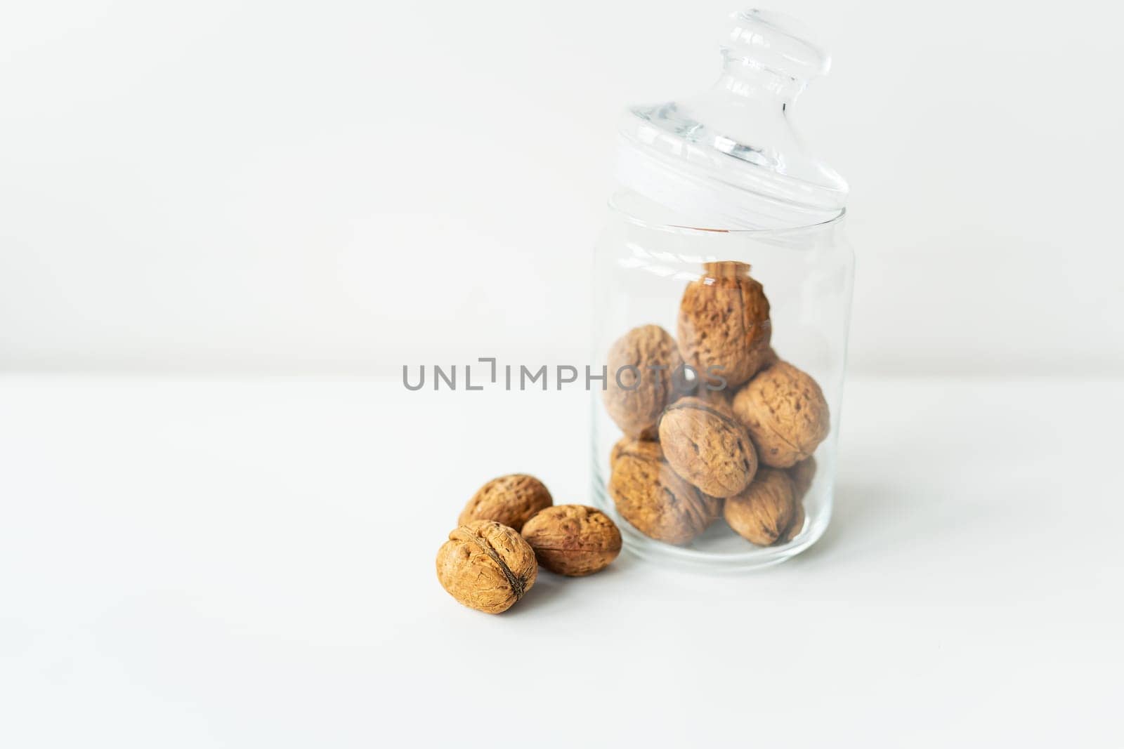 Beautiful whole walnut kernels in a glass jar, whole nuts in the shell can be seen nearby, on a white table. The concept of proper storage of walnuts. Selective focus