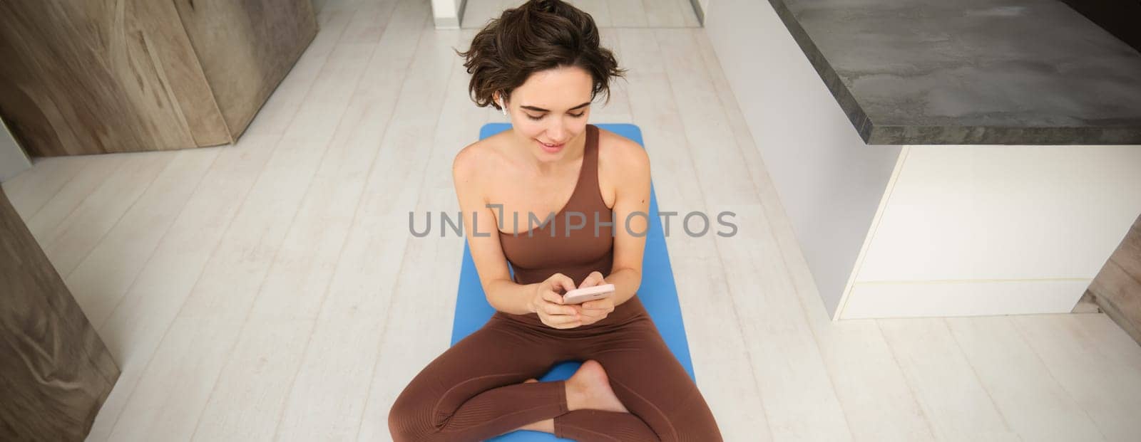 Portrait of fitness girl listens music, workout from home on yoga mat, uses smartphone sport app, watches training session, online exercises.