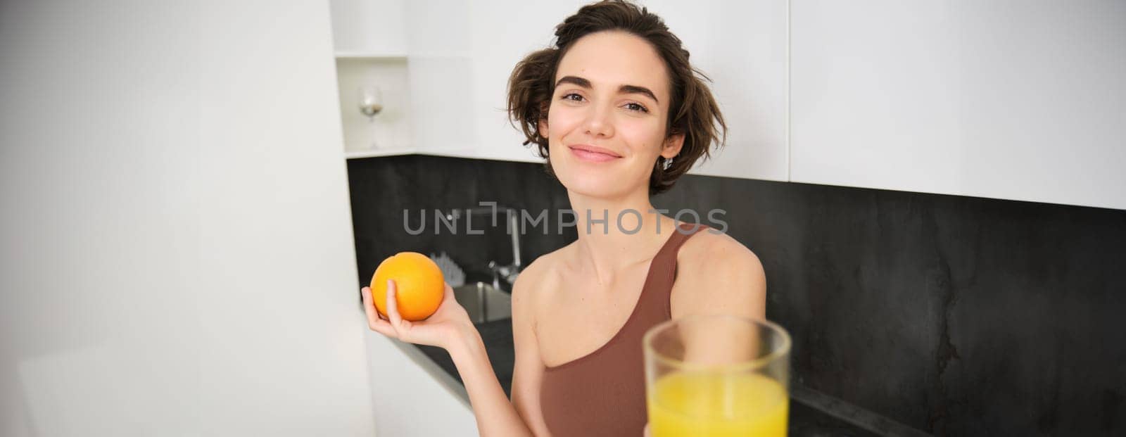 Healthy and beautiful young woman, giving you freshly squeezed glass of juice, holding an orange and smiling, taking vitamin c drink after workout, training and sport activities.