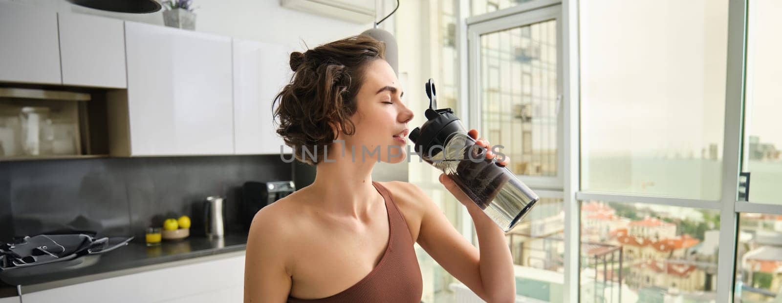 Female athlete in her kitchen, standing with water bottle, drinking after workout yoga training. Sportswear in sports wear stays hydrated after gym exercise.