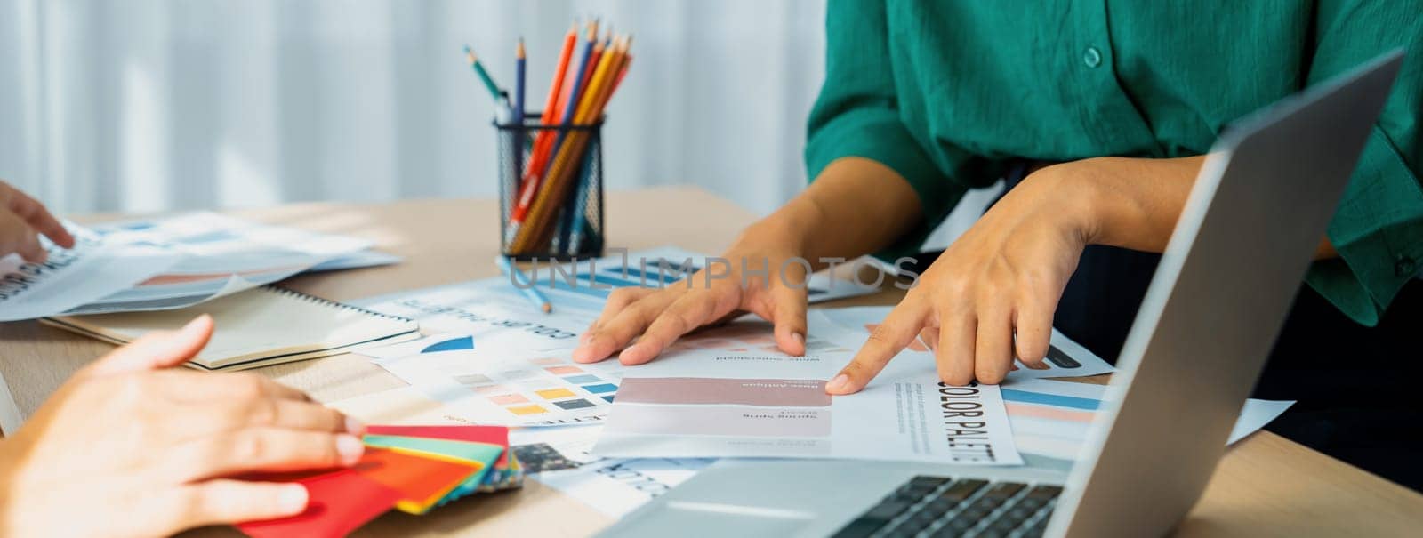 Creative graphic design presents selective color while manager using laptop comparing suitable color at table with color palettes, graphic material scatter around at modern office. Variegated.