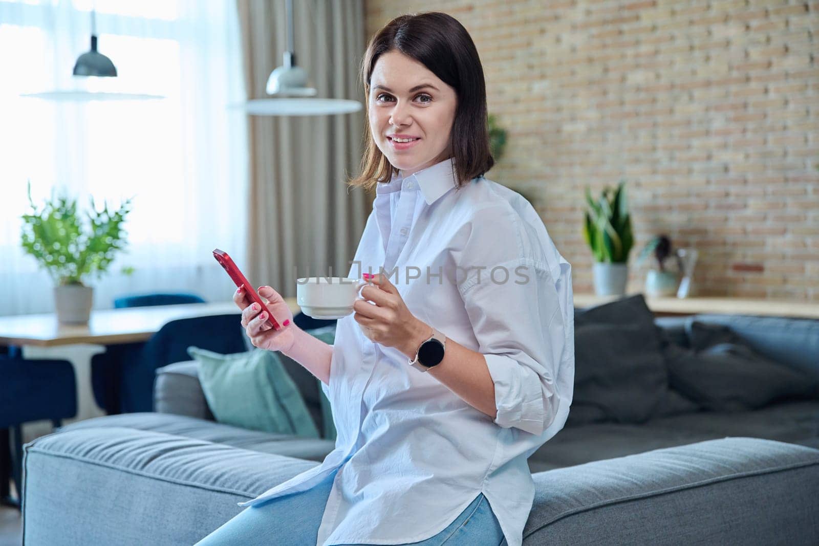Young fashionable beautiful woman at home on couch in living room using smartphone, with cup of coffee in hands. Internet online technology, mobile applications for study leisure life work shopping