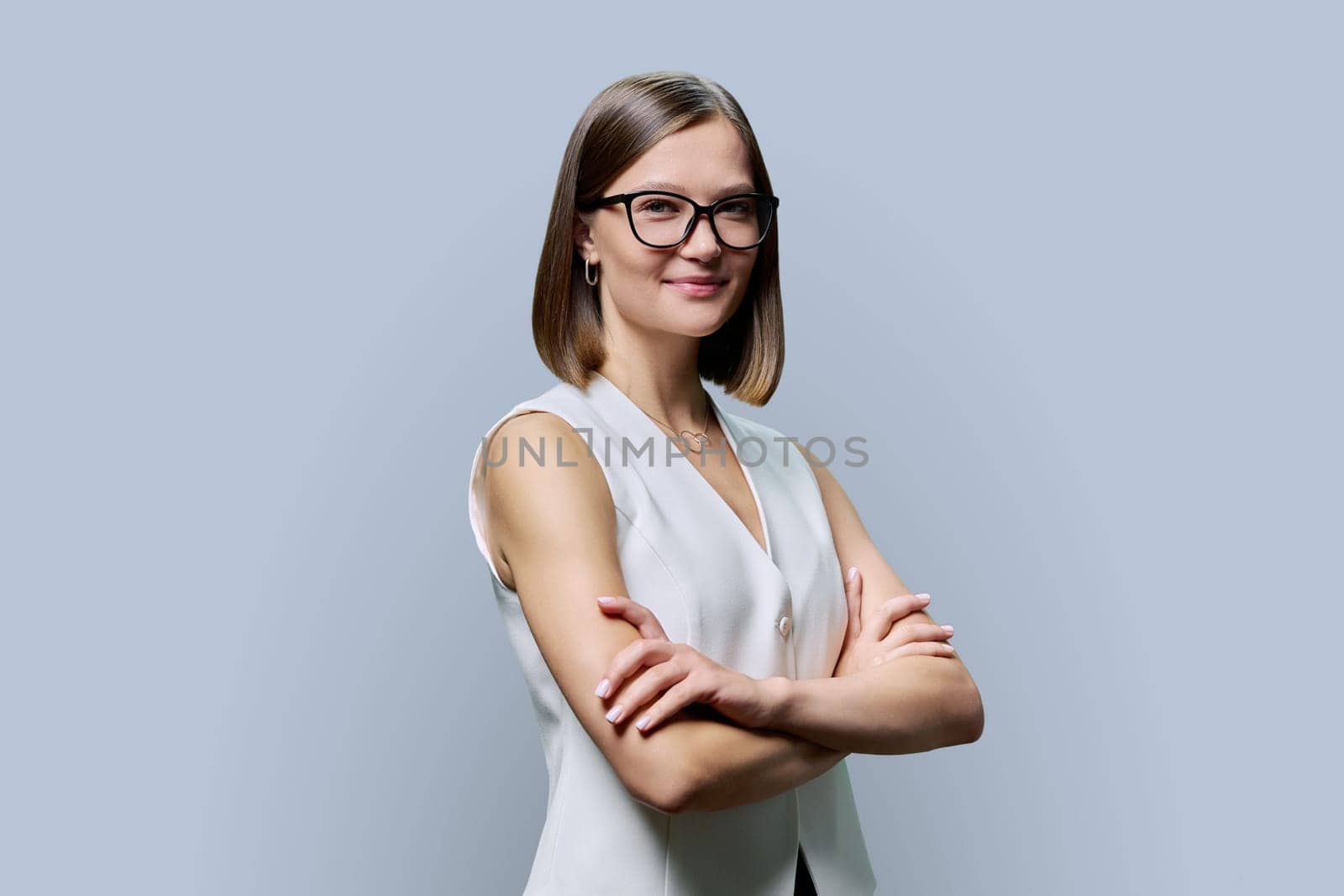 Young smiling woman in glasses with crossed arms posing on gray studio background. Positive business confident successful female looking at camera with folded hands. Business, work, training, people