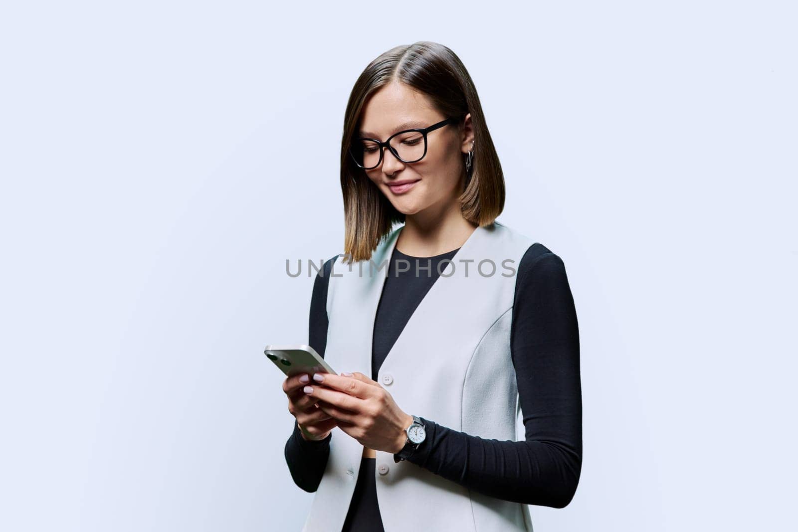 Young confident woman with smartphone in hands on white studio background. Serious female in glasses looking at phone screen texting reading. Using mobile applications for work business study leisure
