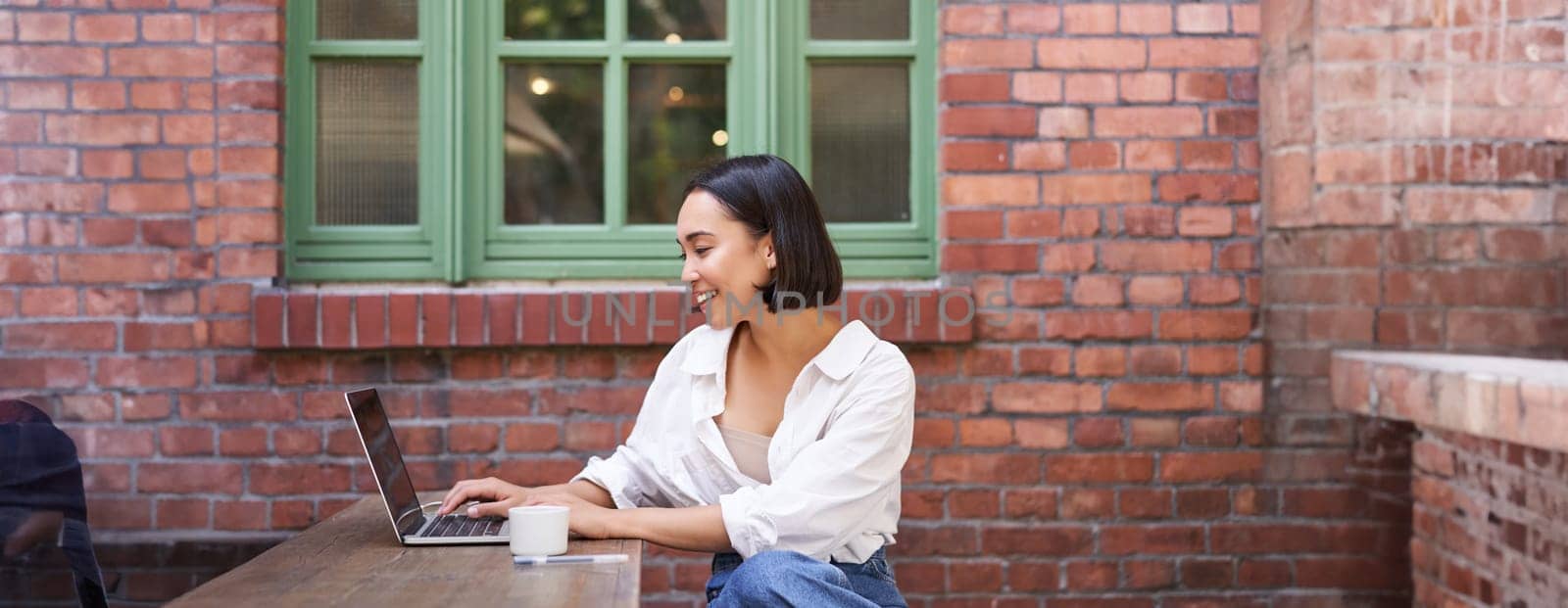 Portrait of young stylish woman, influencer sitting in cafe with cup of coffee and laptop, smiling and looking confident.