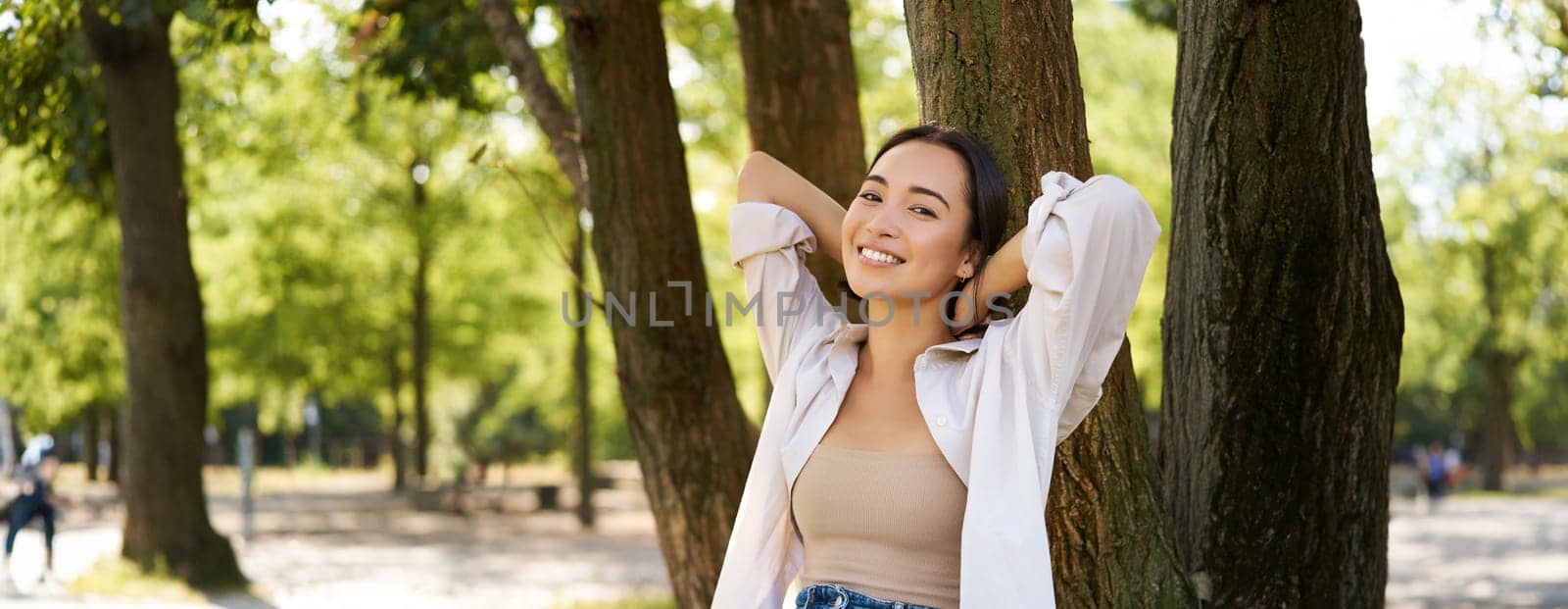 Portrait of beautiful asian woman resting near tree, relaxing in park, smiling and looking happy. Copy space