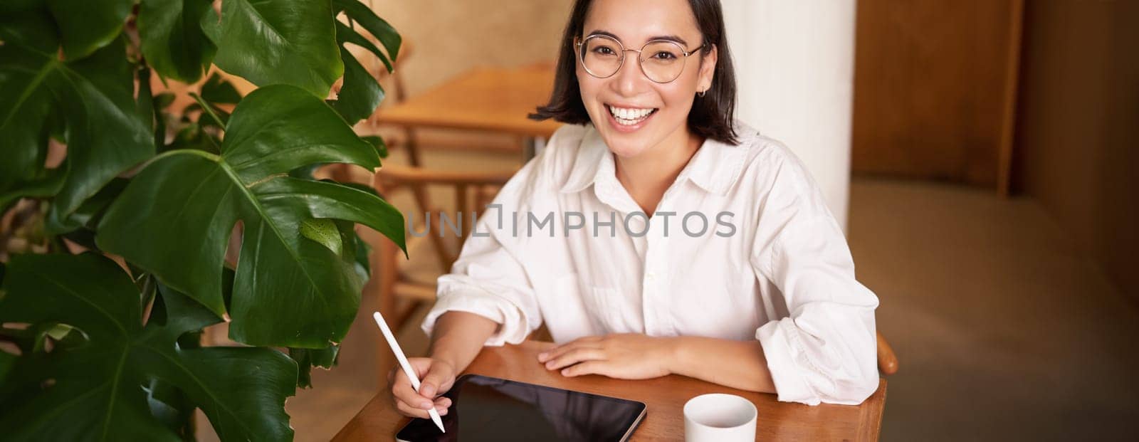 Smiling young female creator, creative girl drawing on digital tablet with graphic pen, drinking coffee in cafe.