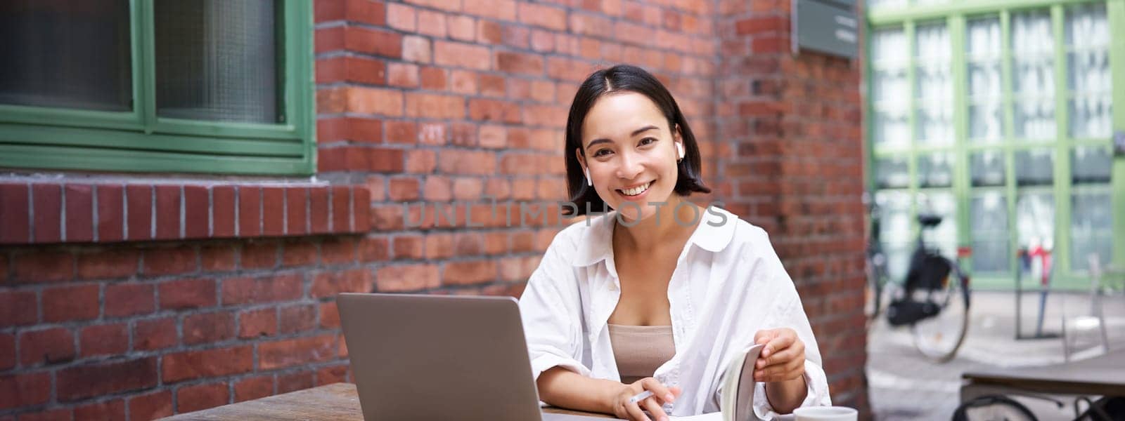 Working woman sitting in coworking space, drinking coffee and using laptop, wearing wireless headphones, watching video and smiling.