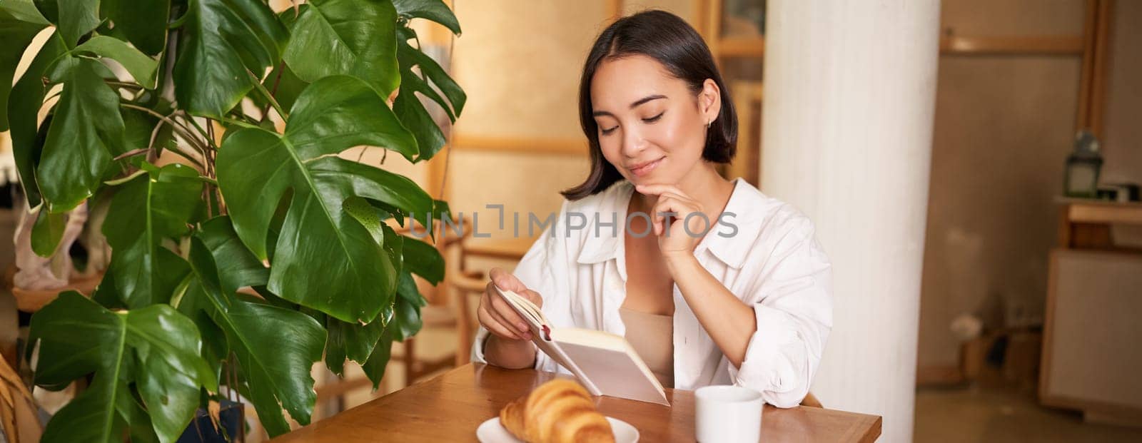 Smiling beautiful girl with a book, reading with thoughtful smirk, thinking of something, drinking coffee in cafe.