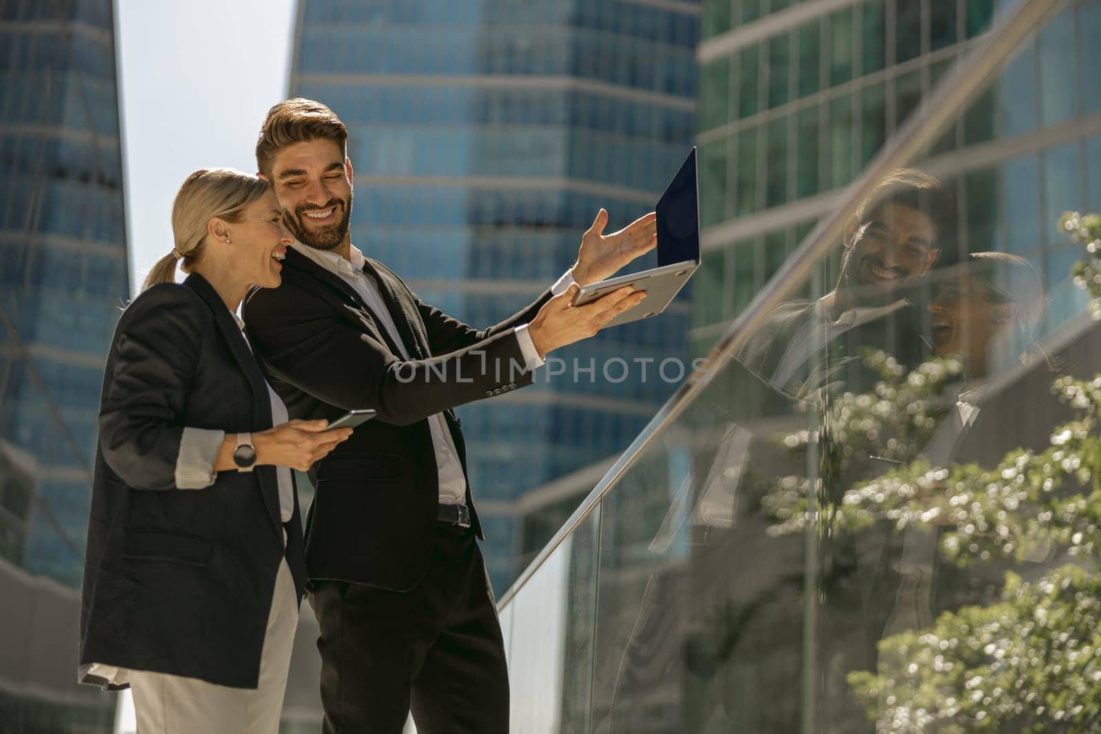 Man and woman in classic suit discussing business details and using laptop while standing outdoors by Yaroslav_astakhov