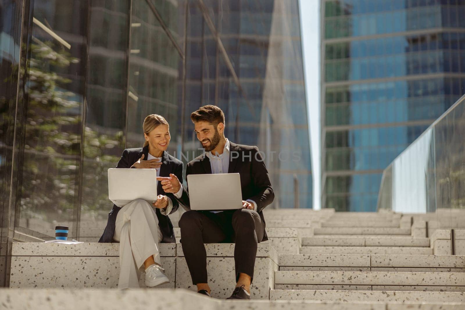 Man and woman in classic suit discussing business details and using laptop while sitting outdoors