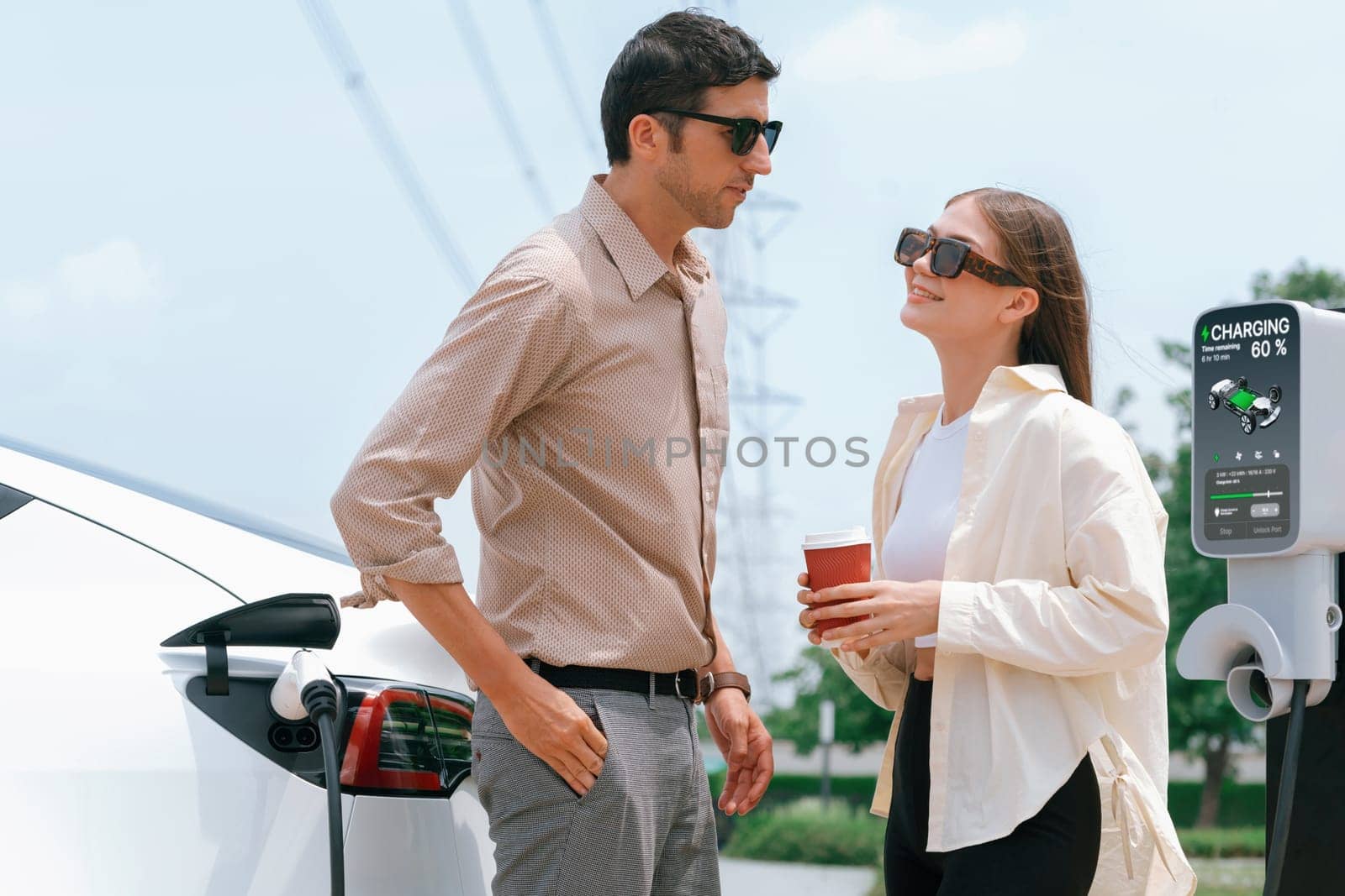 Young couple with coffee recharge EV car battery at charging station connected to electrical industrial power grid tower. Couple with shopping and travel using eco electric car lifestyle. Expedient