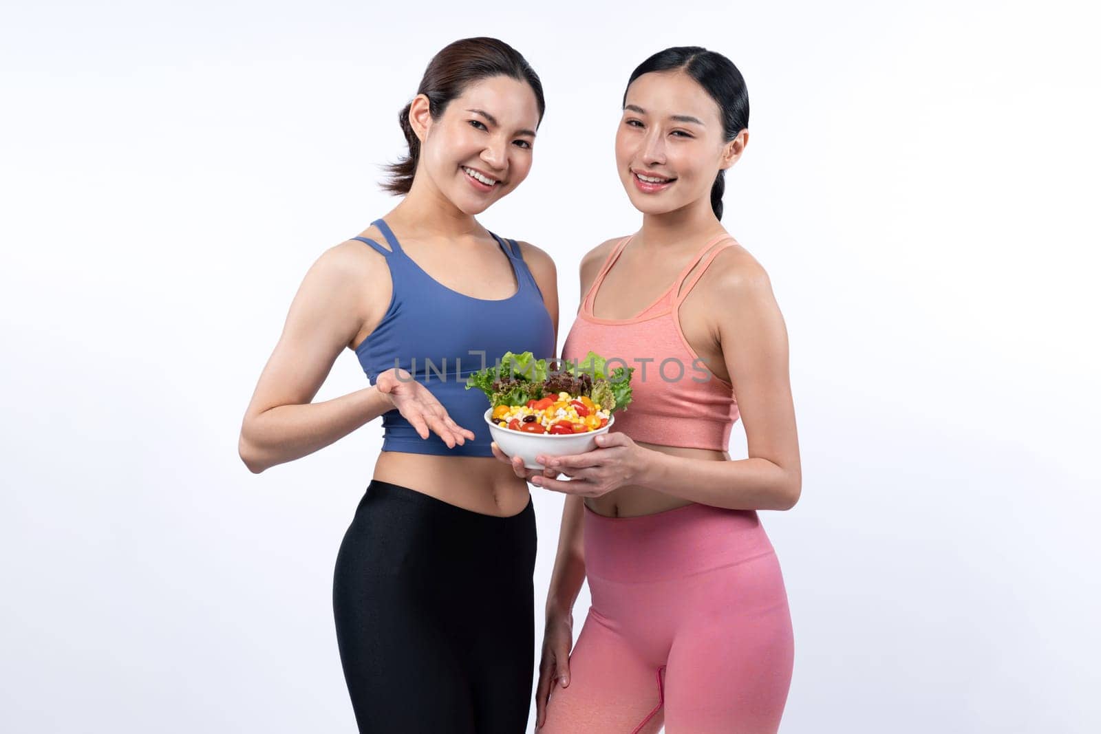 Two young sporty Asian women in sportswear holding salad bowl fill with fruit and vegetable. Natural youthful and fit body lifestyle people with balance nutrition on isolated background. Vigorous