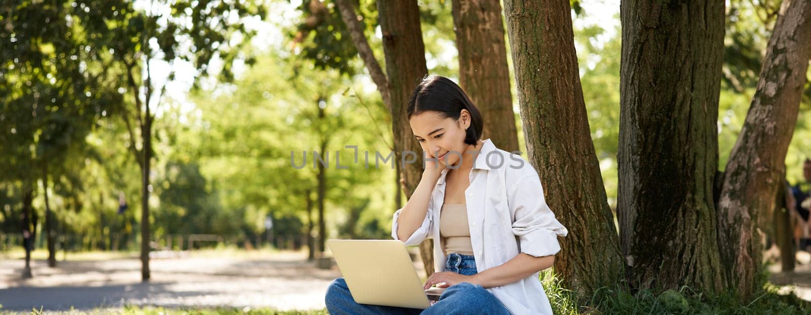 Happy young asian girl sits in park near tree, looking at laptop, working remotely from outdoors, talking to someone, video chat.