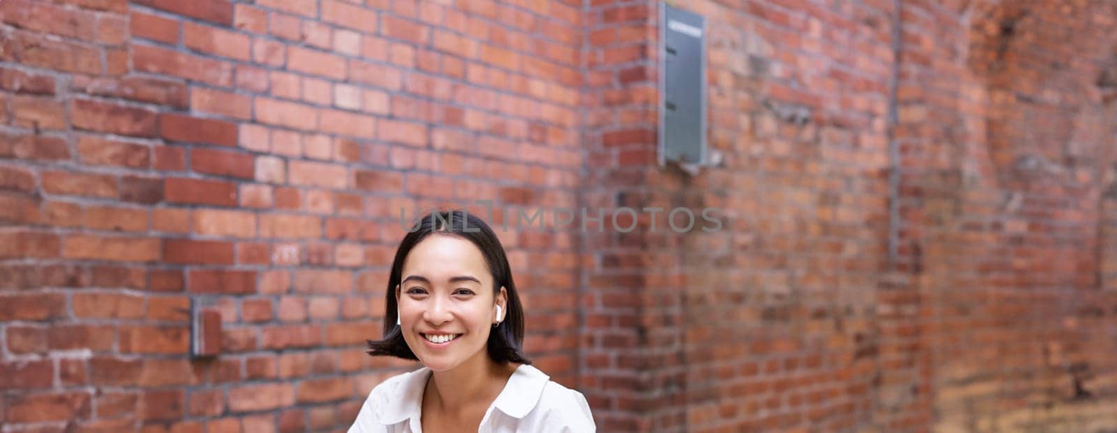 Stylish asian girl works in coworking with laptop, makes notes, uses wireless earphones and smiles, sits near brick wall in office by Benzoix