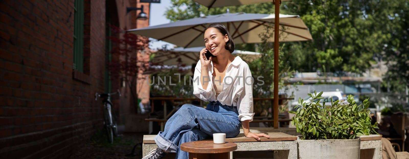 People and communication. Stylish asian girl sits in outdoor cafe with cup of coffee and smartphone, using mobile phone by Benzoix