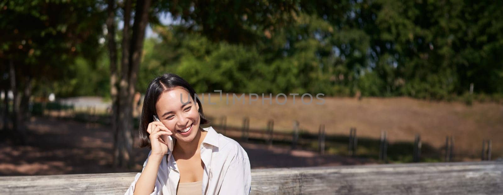 Smiling asian girl relaxing in park, talking on smartphone, having a mobile call while resting outdoors.