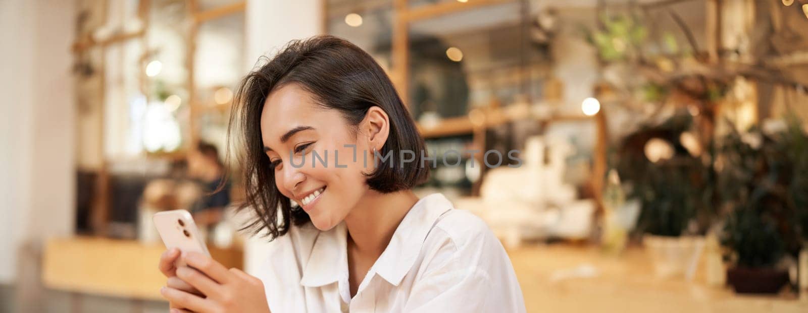 Vertical shot of brunette asian girl sitting in cafe, using smartphone app, chatting on mobile phone and smiling by Benzoix