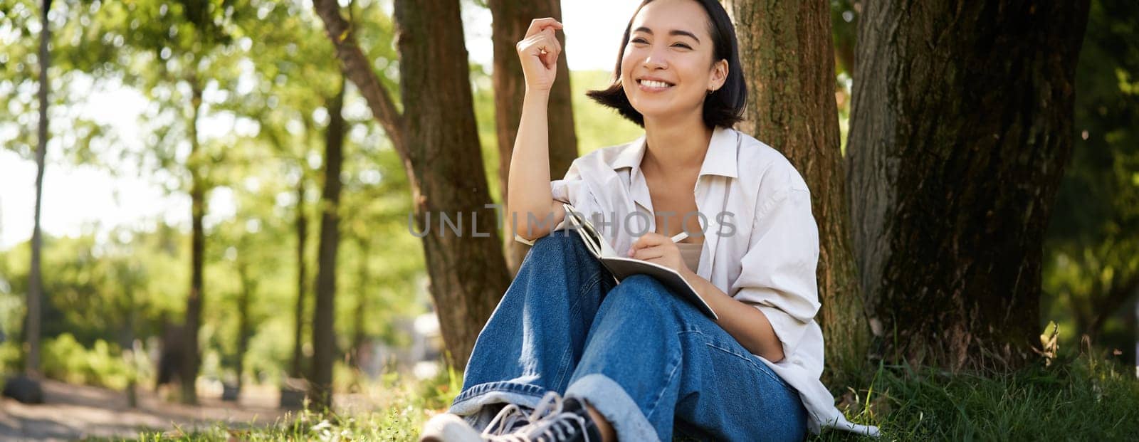 Portrait of young asian woman writing in her notebook, expressing her thoughts on paper in diary, smiling and sitting under tree on summer day in park.