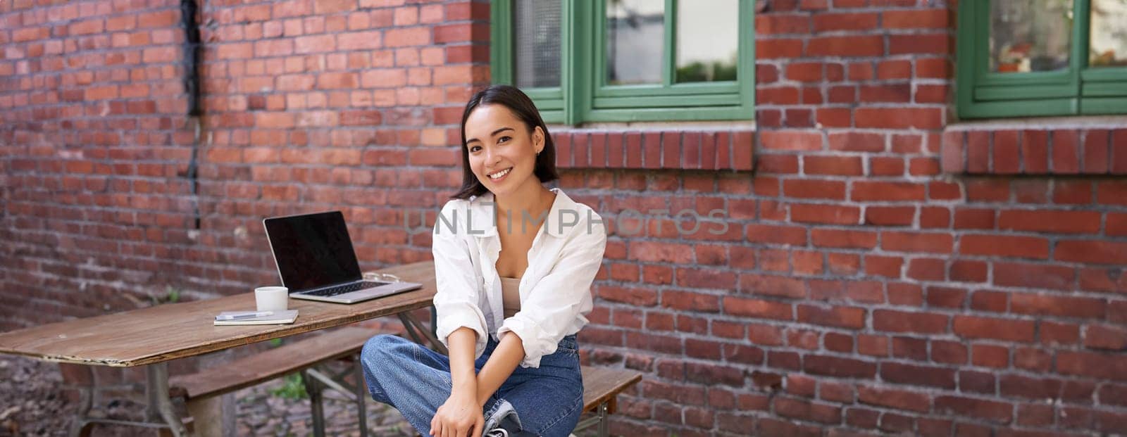 Stylish young businesswoman, asian girl with laptop, sitting in outdoor cafe with cup of coffee and working, using computer.