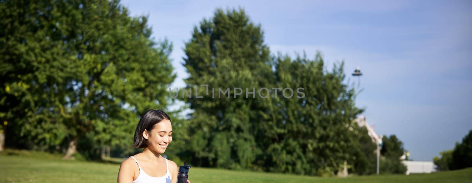 Smiling young asian woman sits on grass, doing exercises in park, workout on lawn, drinks water from bottle.