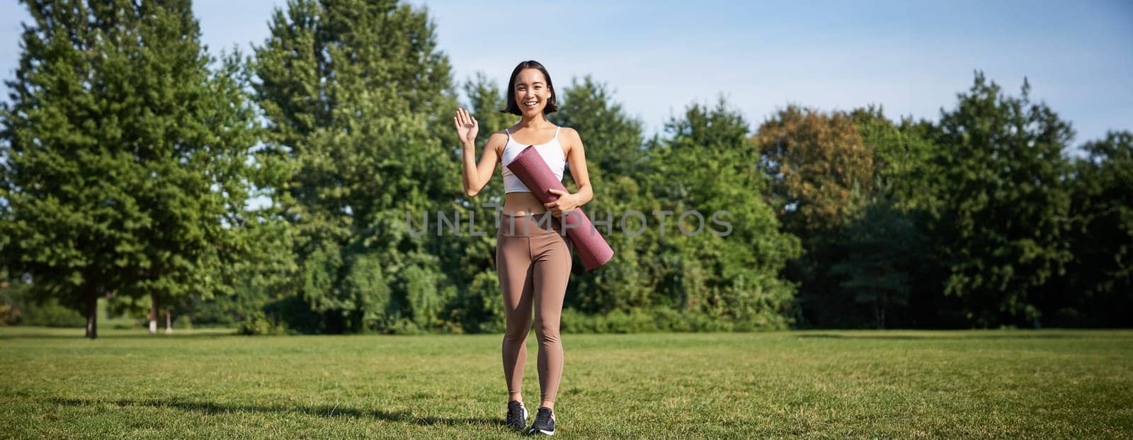 Stylish sportswoman with rubber mat for yoga, says hello, waves at you, ready for workout training outdoors in park on lawn.