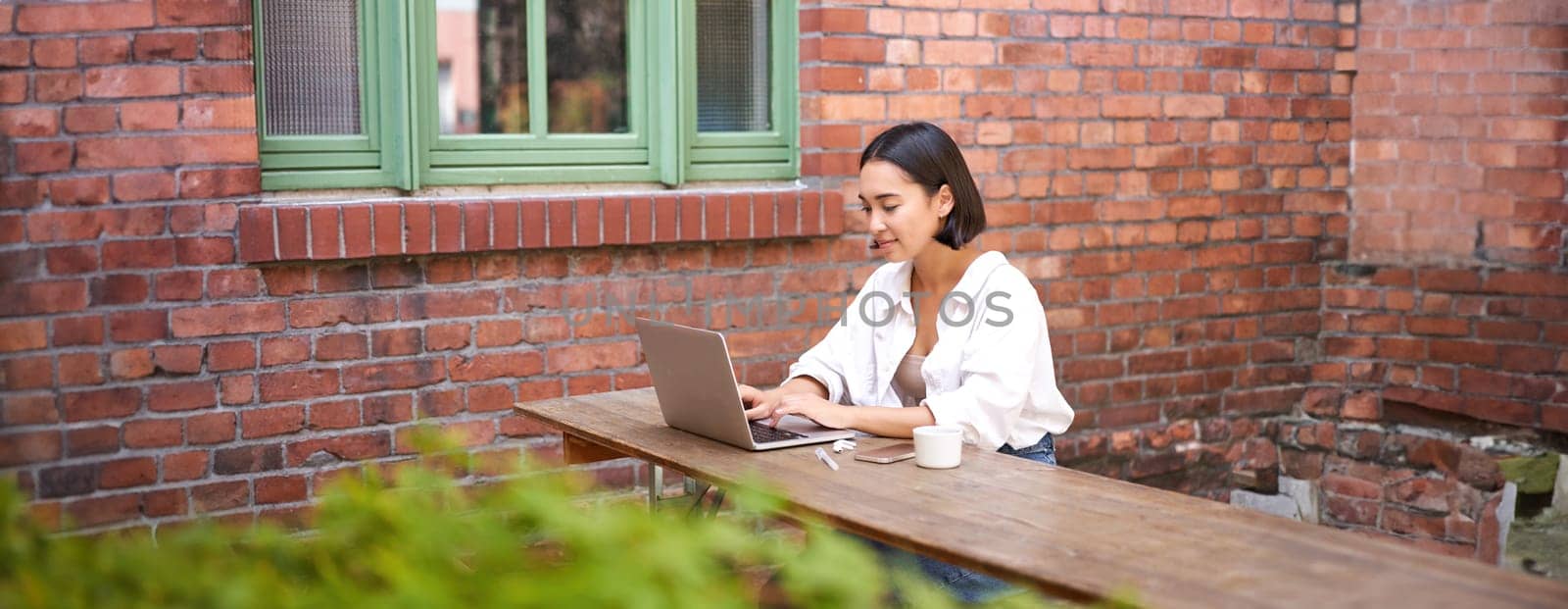 Stylish young urban girl in outdoor cafe, sitting on bench with laptop, smiling, browing on computer by Benzoix