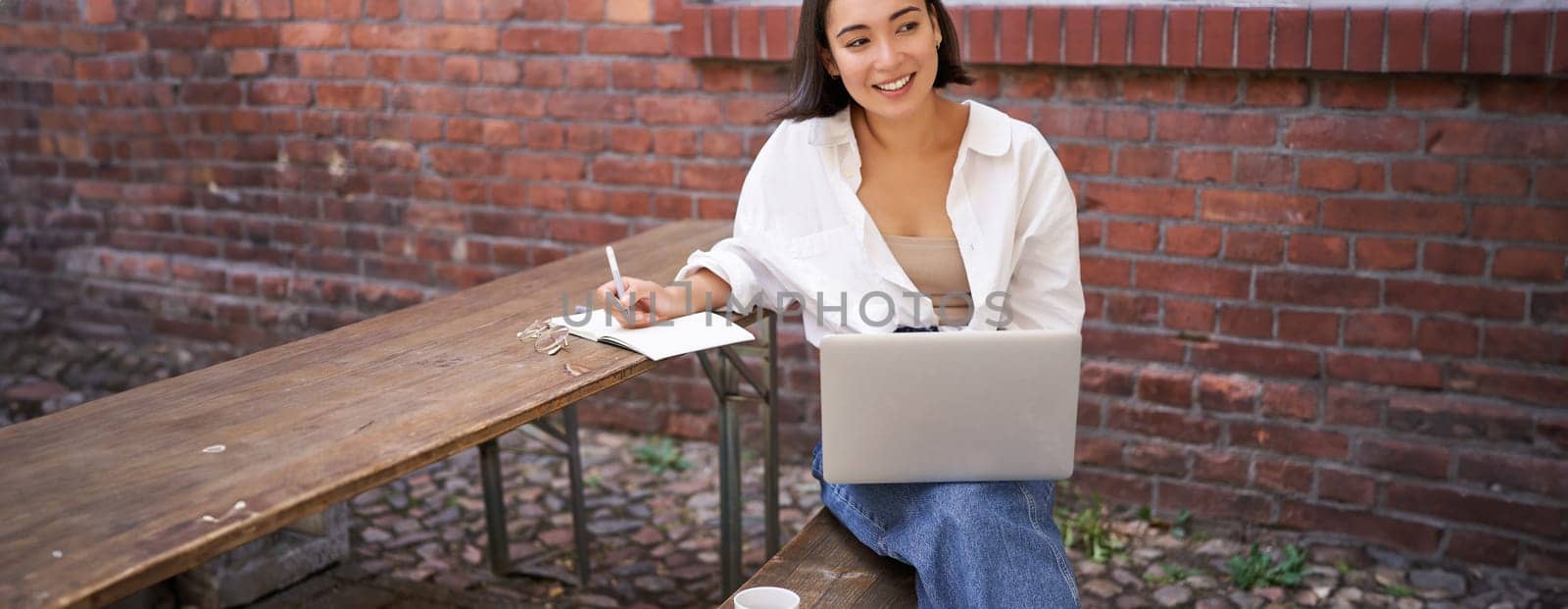 Working woman sitting outdoors in cafe, looking at laptop and making notes in notebook, writing down while watching smth on computer by Benzoix