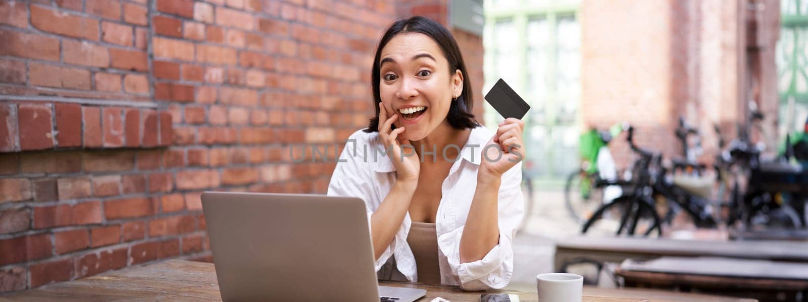 Portrait of young stylish girl sits near laptop, shows credit card and smiles, pays bills, shops online. shopping and mobile banking concept