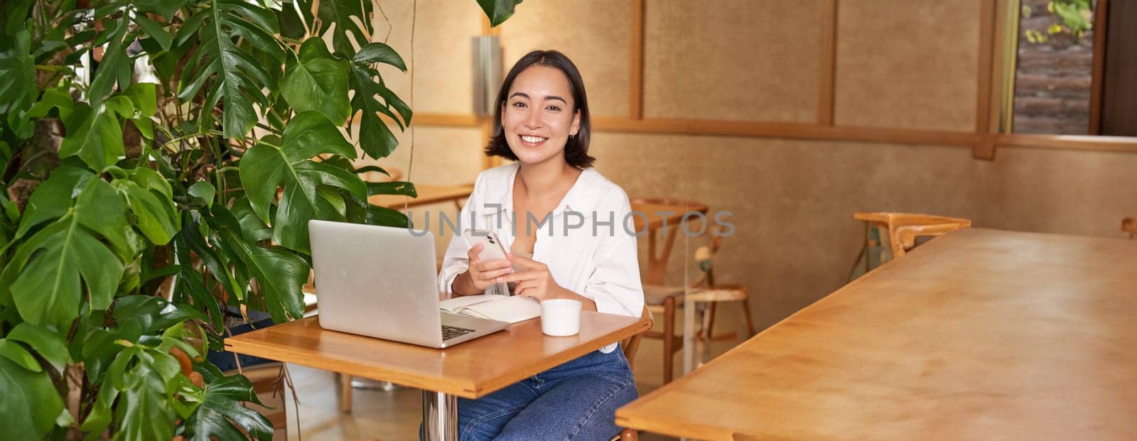 Vertical shot of young asian girl sits in cafe with laptop and smartphone, relaxing and surfing the net, working remotely.
