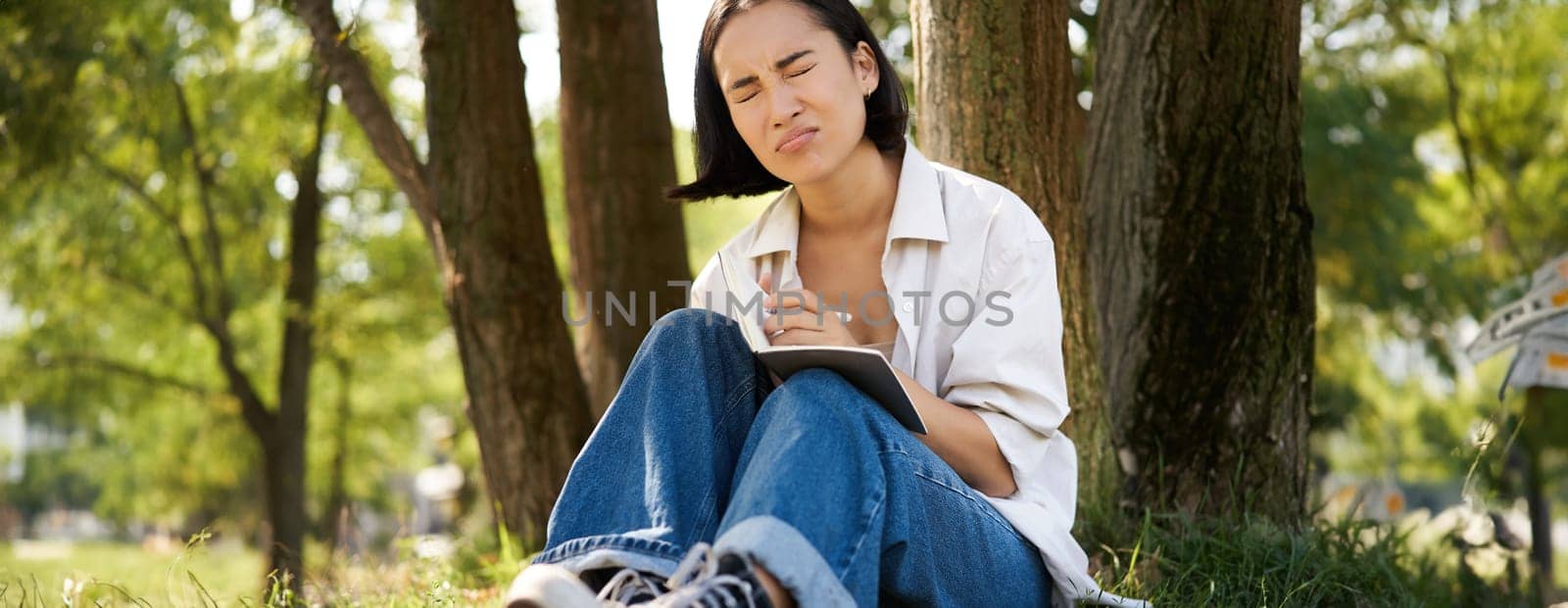 Portrait of sad asian girl writing in her diary and feeling uneasy, sitting in park alone under tree, expressing her distress in notebook.