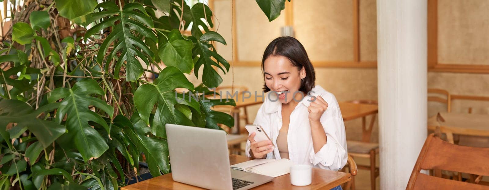 Joyful young asian woman winning, celebrating victory or achievement, receive good news on mobile phone, sitting with laptop and working.