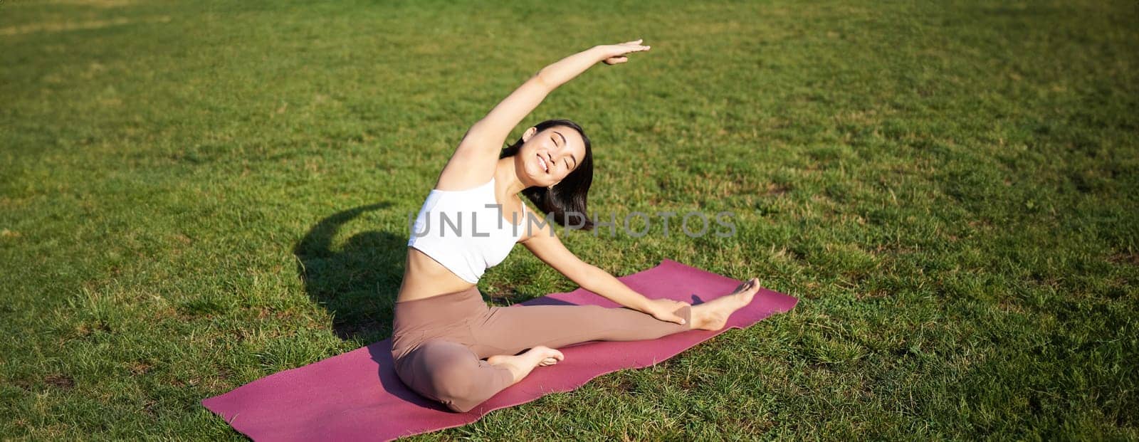 Young woman does yoga on lawn in park, stretching on fitness mat, wellbeing concept.