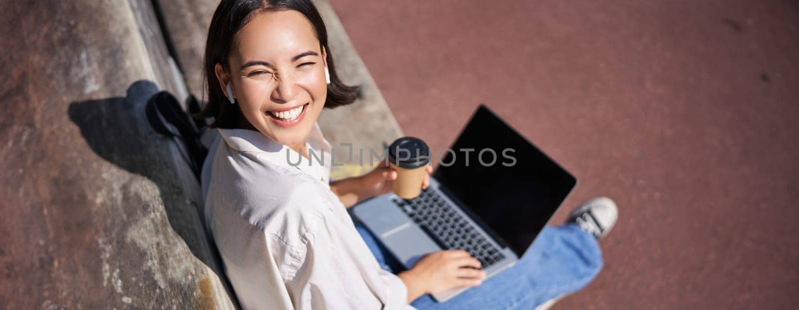 Beautiful young asian female student, sitting with laptop on bench in park, drinking takeaway coffee and listening music, working remotely, studying outdoors.