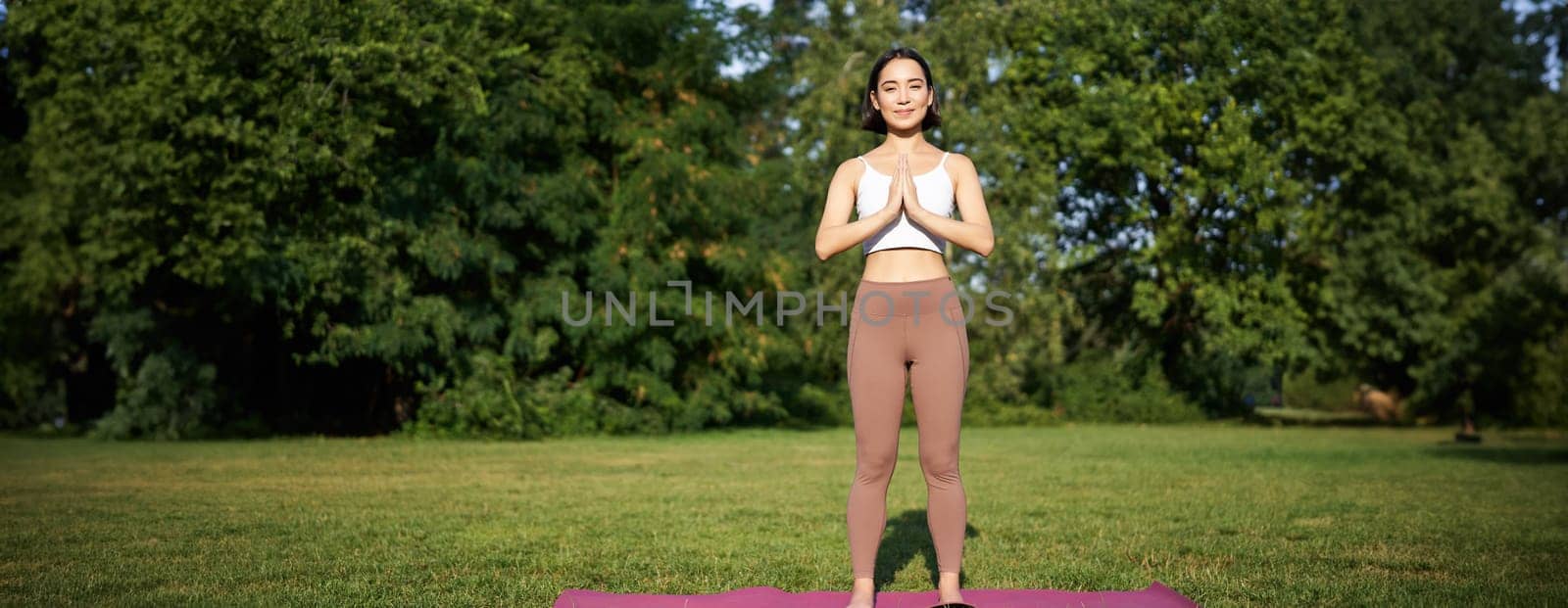 Young mindful woman, meditating on fresh air, doing yoga exercises in park, standing on rubber mat in sportswear.