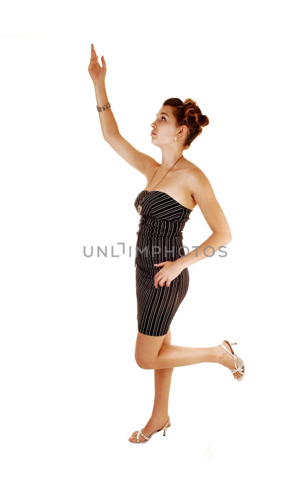 A pretty and young woman in a black dress with stripes on standing from the side for white background with one hand the ear,.