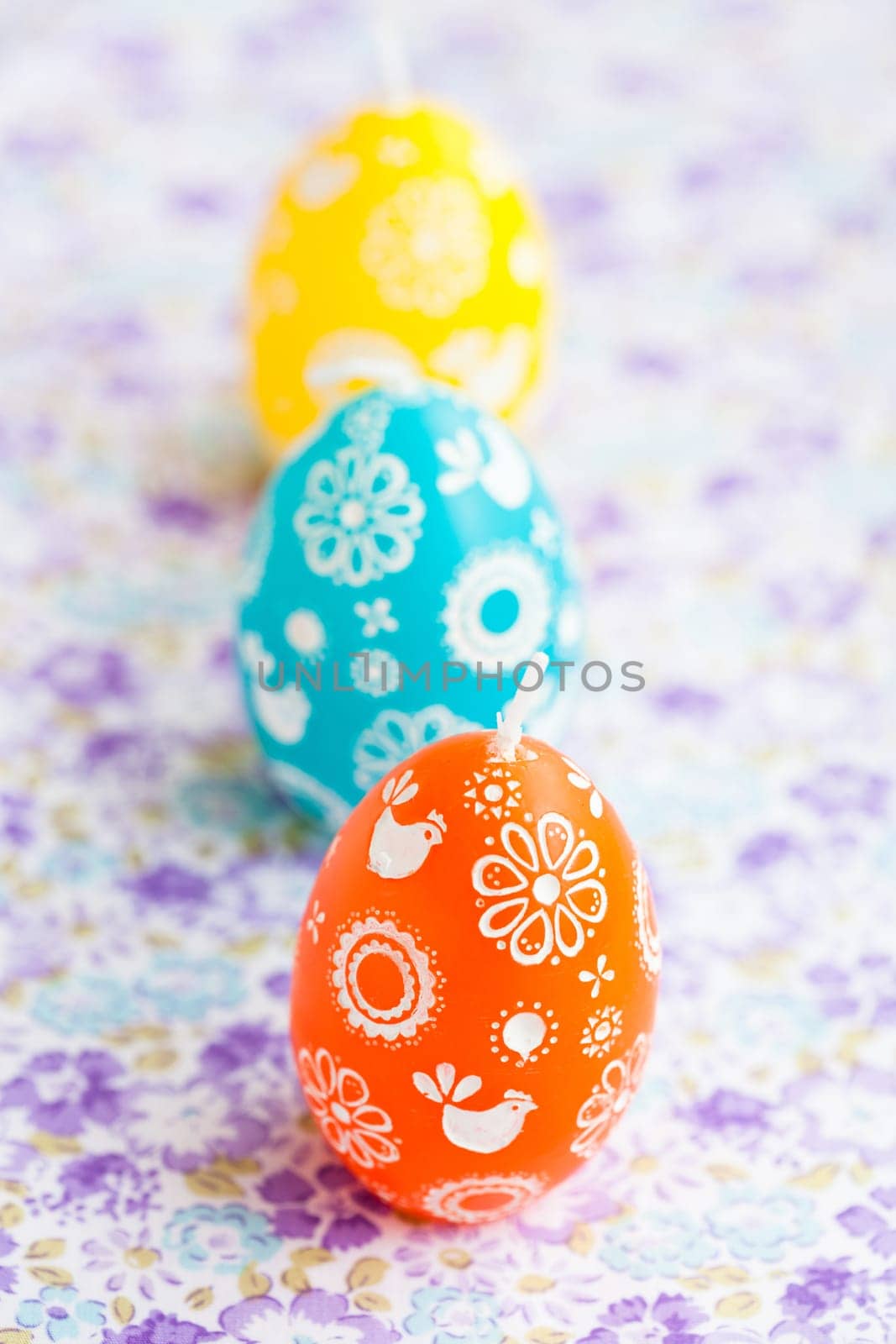 Closeup of three colorful Easter egg shaped candles in row on patterned surface