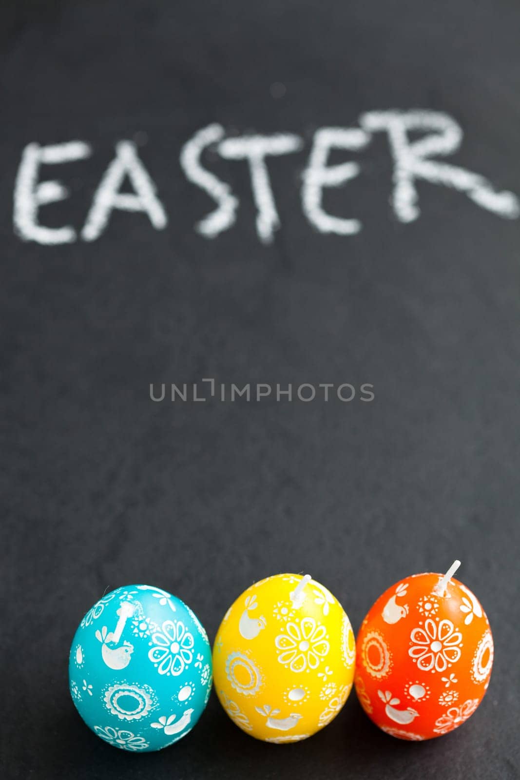 Closeup of three colorful Easter egg shaped candles and text written in chalk on dark background