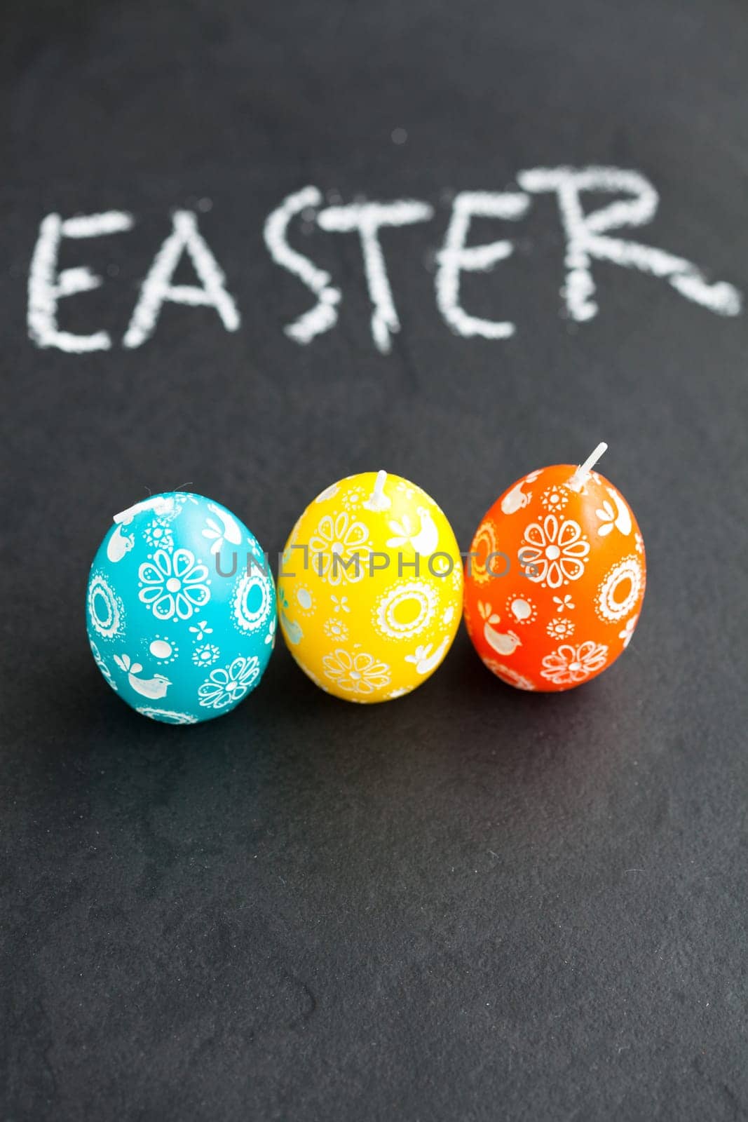 Closeup of three colorful Easter egg shaped candles and text written in chalk on dark background