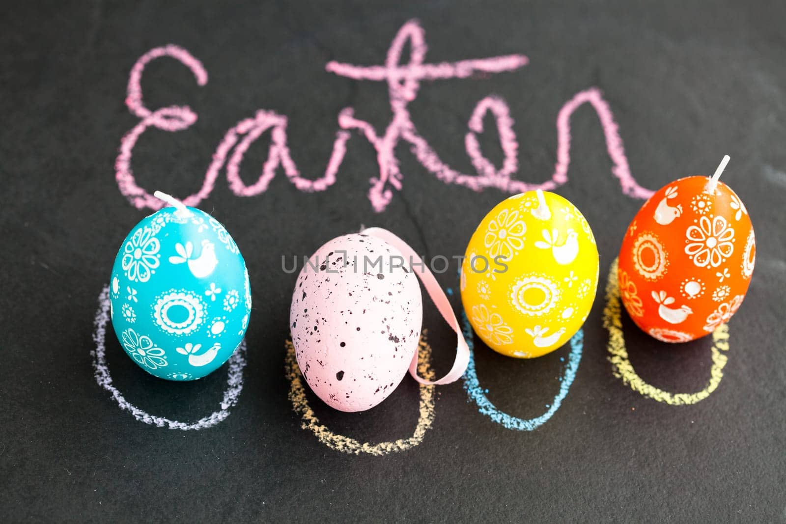 Closeup of four colorful Easter egg shaped candles with Easter text written in pink chalk